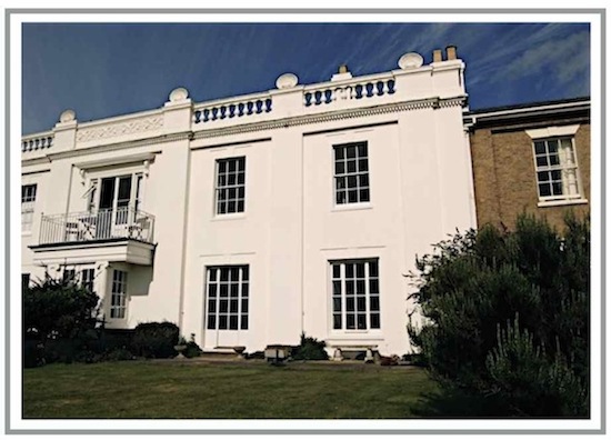 flat white frontage of a what is now a terraced house with four long rectangular windows,  a balustrade edging at the top of the building hiding the roof, with decorative three decorative shells (open clam-shell type) on top.  The terraced house next door has a brick frontage