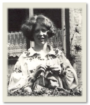 a young woman with thick curly hair cut into a bob, wearing an embroidered smock, looks over from a low hedge knitting.  Behind her is a cottage window with broderie anglaise type net curtain